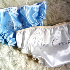 May include: Two pairs of satin panties, one light blue and one white, with ruffled edges. The blue pair says "Bride" in silver glitter, and the white pair says "Mrs. Cruise". Both have a small bow.