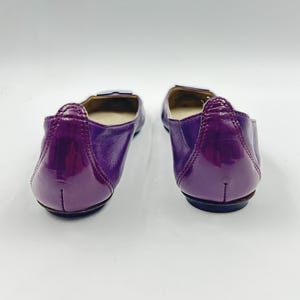 Op de afbeelding: Een paar paarse ballerina's met een glanzende afwerking. De schoenen hebben een ronde neus en een kleine, decoratieve gesp. De achterkant van de schoen heeft een glanzende lakleren uitstraling.