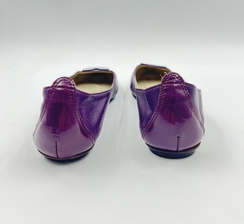 Op de afbeelding: Een paar paarse ballerina's met een glanzende afwerking. De schoenen hebben een ronde neus en een kleine, decoratieve gesp. De achterkant van de schoen heeft een glanzende lakleren uitstraling.