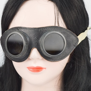 Könnte beinhalten: Schwarze Lederbrille mit runden, dunklen Gläsern und einem beigefarbenen Band. Die Brille wird von einer Puppe mit schwarzen Haaren getragen.