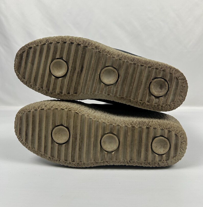 May include: Close-up of the soles of two shoes. The soles are made of a light brown material with a ridged pattern and three circular indentations. The shoes appear worn.