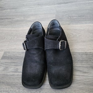 K&ouml;nnte beinhalten: Ein Paar schwarze Wildlederschuhe mit Schnallenverschluss. Die Schuhe haben eine quadratische Spitze und einen niedrigen Absatz.