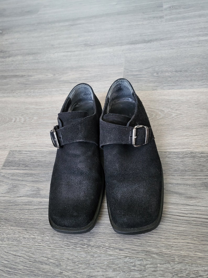K&ouml;nnte beinhalten: Ein Paar schwarze Wildlederschuhe mit Schnallenverschluss. Die Schuhe haben eine quadratische Spitze und einen niedrigen Absatz.