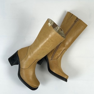 Puede incluir: Un par de botas de cuero color canela con tacones y suelas negros. Las botas tienen ca&ntilde;a alta, cremallera lateral y punta cuadrada. El forro interior es visible en la parte superior de las botas.
