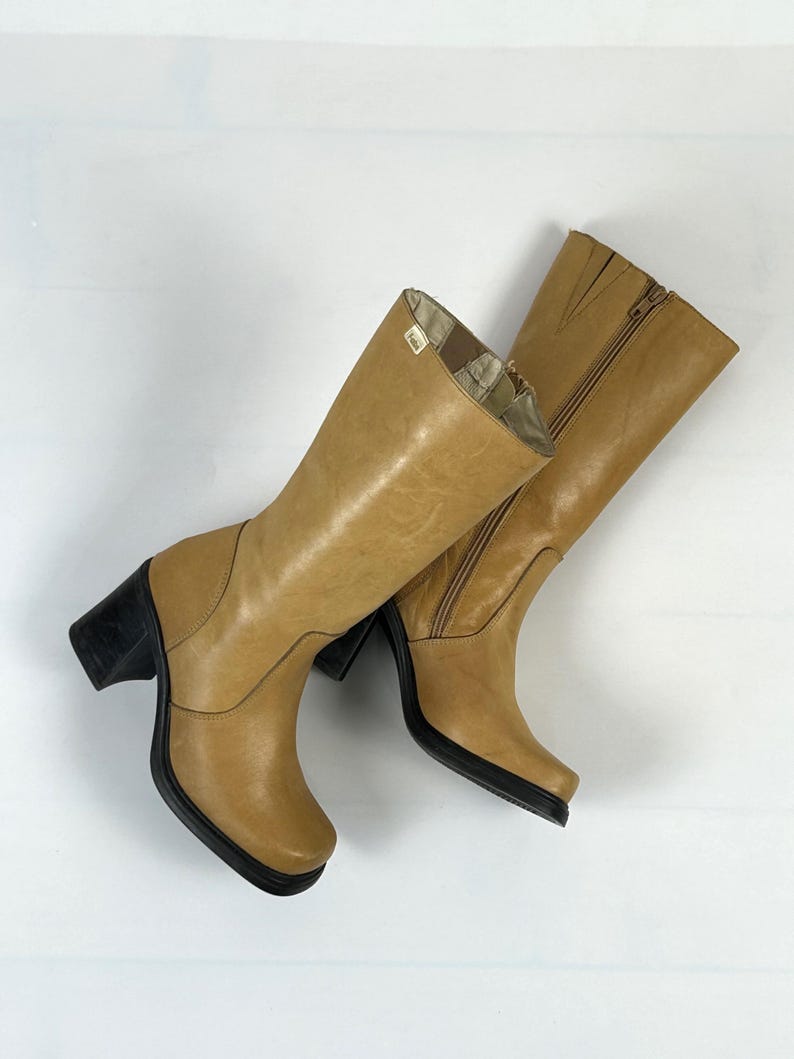 Puede incluir: Un par de botas de cuero color canela con tacones y suelas negros. Las botas tienen ca&ntilde;a alta, cremallera lateral y punta cuadrada. El forro interior es visible en la parte superior de las botas.