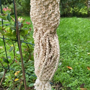 Può includere: Un portavaso in macramè beige con un design intrecciato. Il portavaso è sospeso da un anello e ha un fondo annodato e sfrangiato. Lo sfondo presenta erba verde e fogliame.