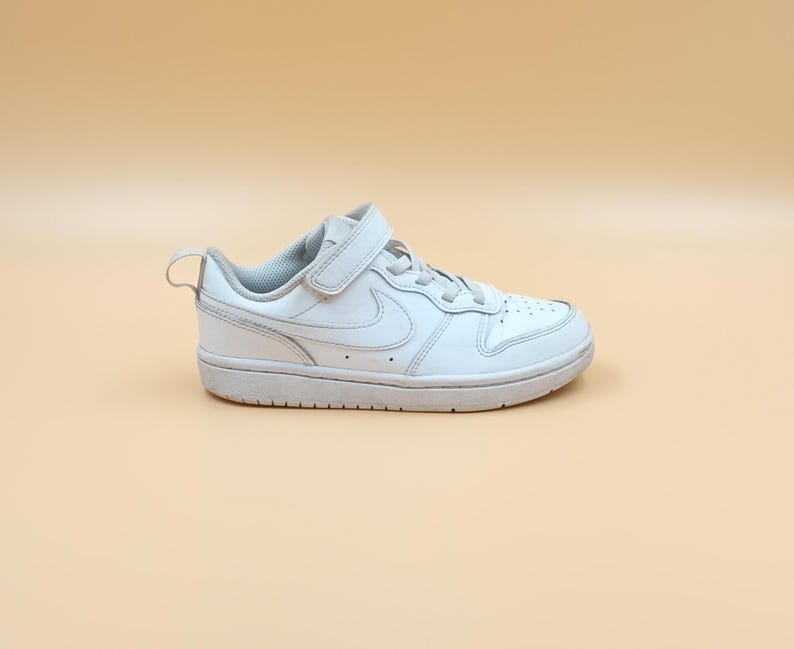 以下が含まれることがあります： ベルクロストラップとナイキのスウッシュロゴが付いた白い革のスニーカー