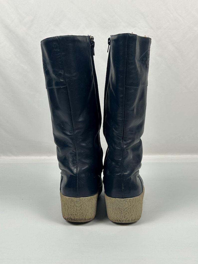 May include: A pair of navy blue leather boots with a beige sole. The boots have a zipper on the side and a textured pattern near the top. The boots are approximately 12 inches tall.