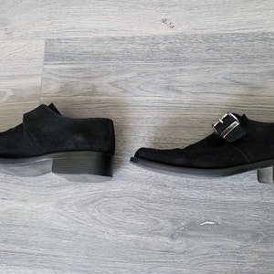 K&ouml;nnte beinhalten: Ein Paar schwarze Wildlederschuhe mit Schnallenverschluss. Die Schuhe haben einen niedrigen Absatz und eine spitze Zehenpartie.