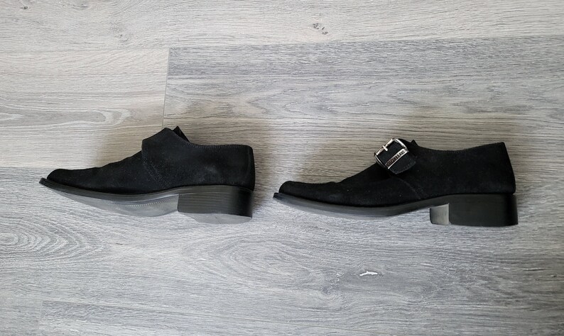 K&ouml;nnte beinhalten: Ein Paar schwarze Wildlederschuhe mit Schnallenverschluss. Die Schuhe haben einen niedrigen Absatz und eine spitze Zehenpartie.