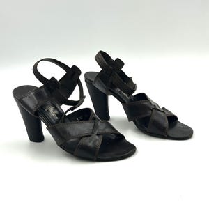 May include: A pair of dark brown leather sandals with a crisscross design over the toes and ankle straps. The sandals have thick, rounded heels. The background is plain white.