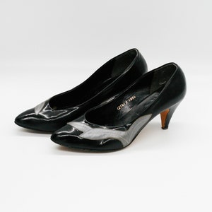 May include: A pair of black leather pumps with a pointed toe and a low heel. The shoes have a silver-gray accent on the vamp. The heels are a light brown color.