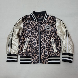 Op de afbeelding: Een bomberjack met luipaardprint met gouden mouwen en zwart-witte gestreepte boorden. De jas heeft een ritssluiting en een kleine ronde patch met tekst. Het woord "KIDS" staat op de kraag.
