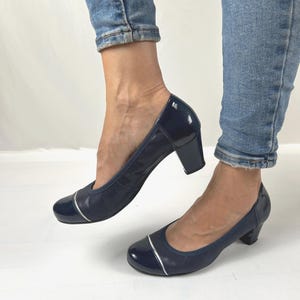 May include: A pair of navy blue pumps with a low block heel. The shoes feature a glossy patent leather toe cap and heel counter, contrasted by a textured fabric side panel. A thin silver metallic accent strip separates the toe cap from the body.