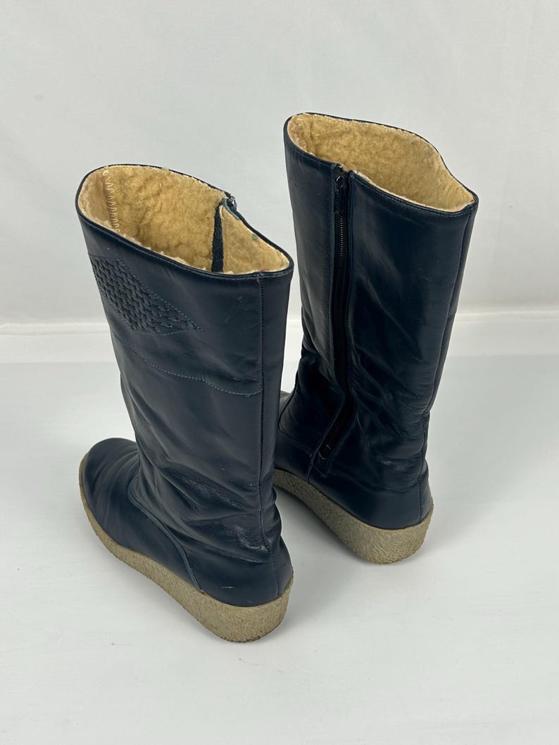 May include: A pair of navy blue winter boots. The boots have a tall shaft with a zipper on the side and a textured design on the upper. The interior is lined with a tan, fuzzy material. The soles are a light beige color.