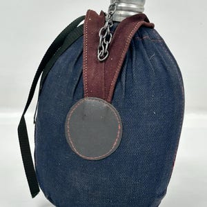 Può includere: Una borraccia blu navy con tappo e catena argentati, racchiusa in una fodera di tessuto. La fodera presenta un motivo a spina di pesce e un cinturino bordeaux. Sono visibili anche un cinturino nero e una toppa in pelle.