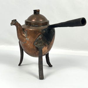 May include: A copper kettle with a rounded body, a lid, and a long, dark handle. The kettle has three legs and a spout that resembles a bird's head. The metal has a patina, suggesting age. The kettle is likely for decorative purposes.