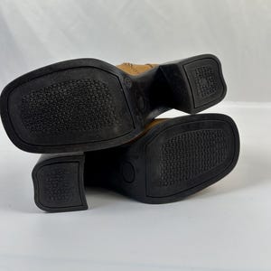 Puede incluir: Un par de botas de cuero color canela con suelas y tacones negros. Las suelas tienen un patr&oacute;n texturizado para el agarre. Las botas tienen punta cuadrada y tac&oacute;n cuadrado. Las botas se muestran de abajo hacia arriba.