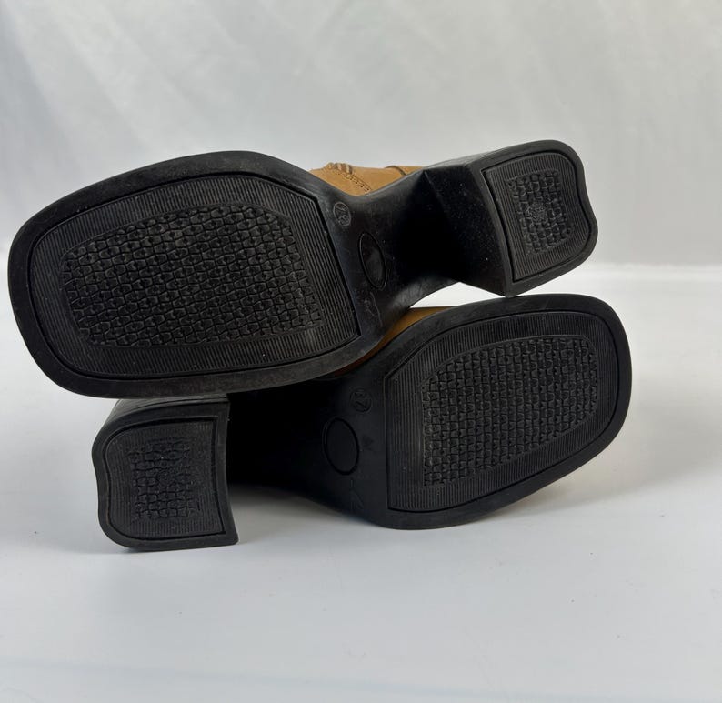 Puede incluir: Un par de botas de cuero color canela con suelas y tacones negros. Las suelas tienen un patr&oacute;n texturizado para el agarre. Las botas tienen punta cuadrada y tac&oacute;n cuadrado. Las botas se muestran de abajo hacia arriba.