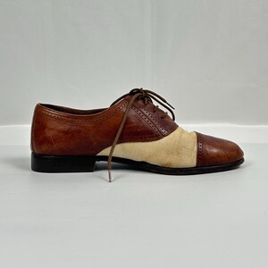 May include: A brown and tan oxford shoe with a black sole. The shoe features a leather upper with a canvas panel on the vamp. The shoe has brown laces and a classic, vintage design.