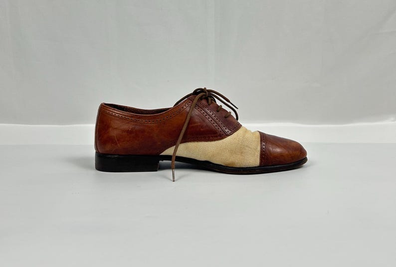 May include: A brown and tan oxford shoe with a black sole. The shoe features a leather upper with a canvas panel on the vamp. The shoe has brown laces and a classic, vintage design.