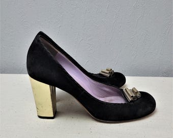 Könnte beinhalten: Schwarze Pumps aus Wildleder mit einem goldenen Blockabsatz und einem silbernen Metallelement an der Spitze.