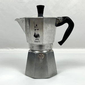 以下が含まれることがあります： 黒いハンドルとつまみが付いた、シルバー色のBialetti Moka Expressエスプレッソメーカー。前面にはBialettiのロゴが見えます。このアルミニウム製エスプレッソメーカーはクラシックなデザインです。