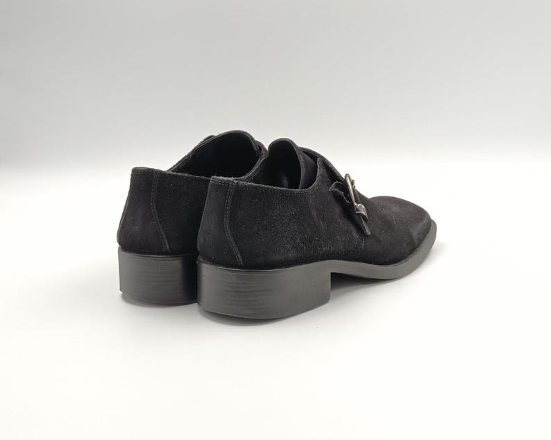 K&ouml;nnte beinhalten: Ein Paar schwarze Schnallenschuhe aus Wildleder. Die Schuhe haben ein klassisches Design mit einer spitzen Zehenpartie und einem niedrigen Absatz.