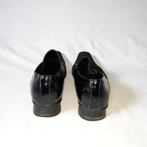 Puede incluir: Un par de zapatos negros de cuero sin cordones con un acabado brillante. Los zapatos tienen una punta redonda y un tac&oacute;n bajo.