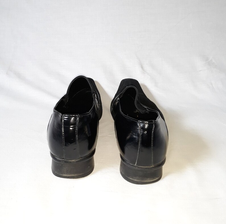 Puede incluir: Un par de zapatos negros de cuero sin cordones con un acabado brillante. Los zapatos tienen una punta redonda y un tac&oacute;n bajo.
