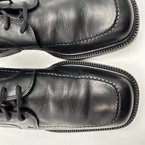 K&ouml;nnte beinhalten: Paar schwarze Lederschuhe mit quadratischen Zehen und Ziern&auml;hten. Die Schuhe haben Schn&uuml;rsenkel und eine strukturierte Sohle. Das Leder weist leichte Knitterfalten auf. Draufsicht.