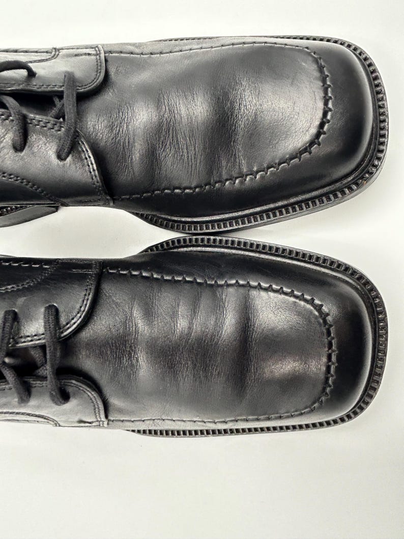 K&ouml;nnte beinhalten: Paar schwarze Lederschuhe mit quadratischen Zehen und Ziern&auml;hten. Die Schuhe haben Schn&uuml;rsenkel und eine strukturierte Sohle. Das Leder weist leichte Knitterfalten auf. Draufsicht.