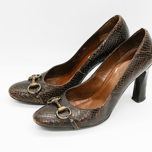 Pode incluir: Um par de sapatos de salto alto de cobra marrom com fivelas de metal dourado. Os saltos são saltos de bloco.