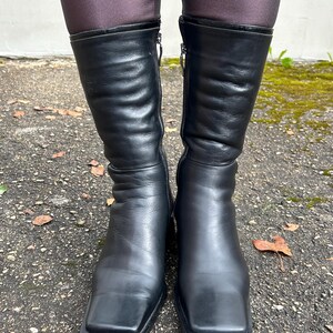 May include: A pair of black leather mid-calf boots featuring a square toe design, a smooth matte finish on the lower foot, and a slightly textured, creased leather shaft. Each boot includes a vertical side zipper with a silver-toned metal pull.