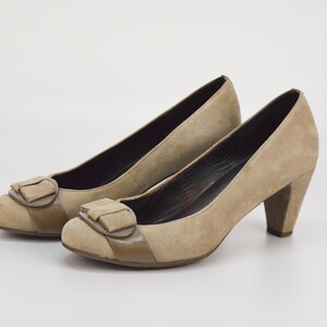 May include: A pair of beige suede court shoes with a rounded toe and a small bow detail on the vamp. The heels are approximately 7.6 cm high. The shoes are set against a white background.