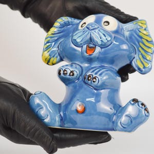 May include: A glossy blue ceramic figurine of a seated dog with large ears featuring yellow accents. The dog has wide white eyes, a black nose, a small orange mouth, and black paw details. A small orange circular accent is visible on the belly.