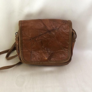 Peut inclure: Un sac bandoulière en cuir marron présentant un motif patchwork avec des coutures visibles sur le rabat avant. Le sac a une forme rectangulaire structurée, une fermeture à rabat et une fine bandoulière en cuir marron assortie sur les côtés.