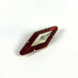 Op de afbeelding: Een vintage broche in ruitvorm. Het heeft een zilverkleurig metalen frame met een rode emaille rand en een centrale, heldere, ruitvormige steen. De broche staat tegen een witte achtergrond.