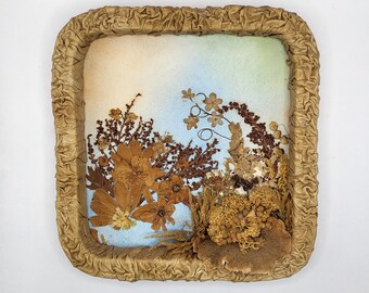 Op de afbeelding: Een vierkant ingelijst kunstwerk van geperste bloemen met een blauwe en witte achtergrond. De bloemen zijn bruin en beige en zijn in het midden van de lijst in een tros gerangschikt.