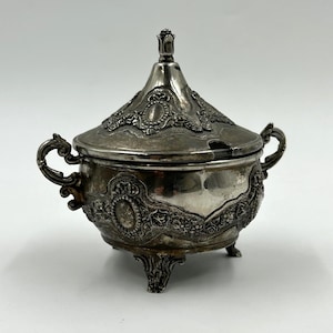 May include: An antique silver sugar bowl with a lid and handles. The bowl is supported by three decorative feet and features detailed floral and scrollwork. The lid is topped with a decorative finial.