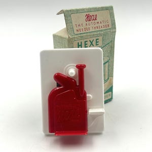 May include: A red Hexe automatic needle threader on a white card, with a green and white box in the background. The box reads "Hexe THE AUTOMATIC NEEDLE THREADER". The threader has a red handle and a white button.