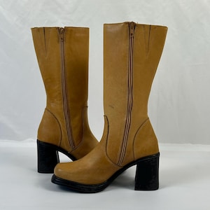 Puede incluir: Un par de botas altas de cuero color canela con tacones de bloque negros. Las botas tienen una cremallera lateral y una puntera redondeada. El cuero tiene un aspecto ligeramente desgastado. Las botas est&aacute;n sobre un fondo blanco.