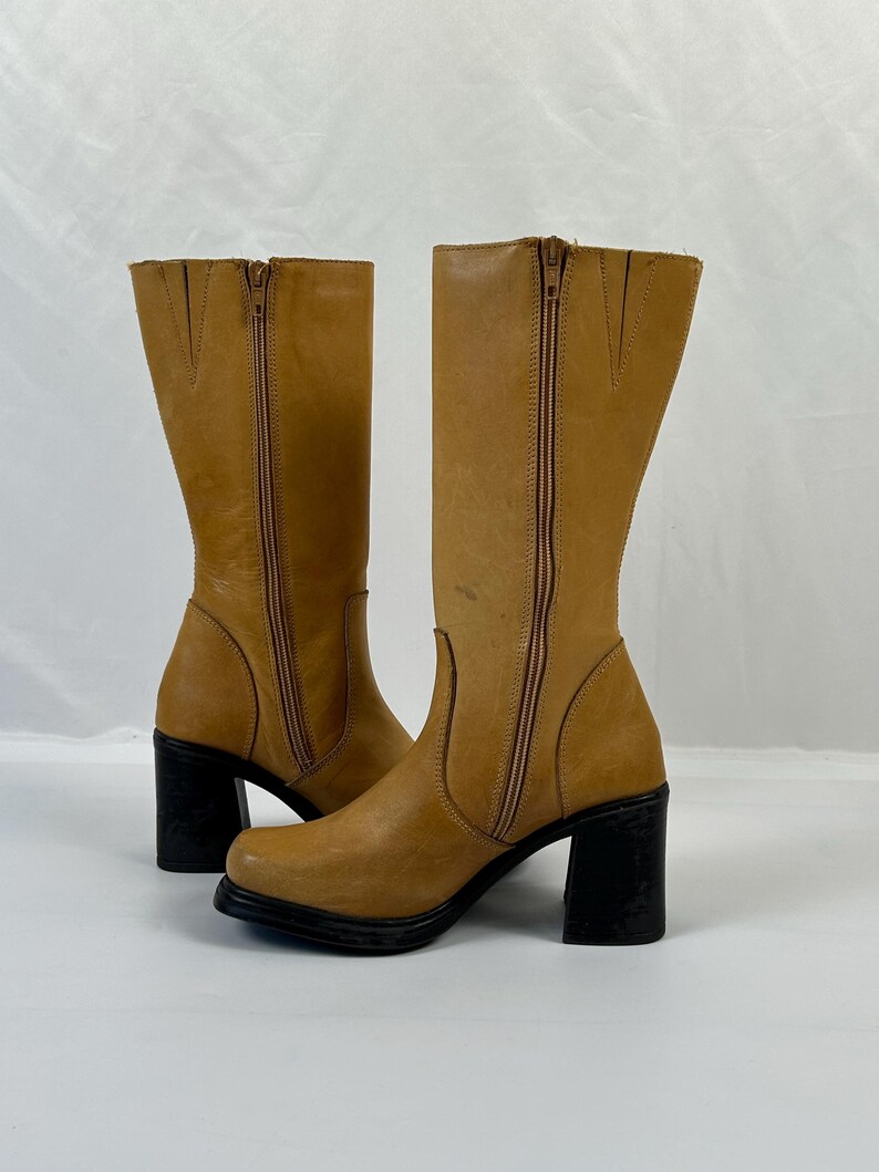 Puede incluir: Un par de botas altas de cuero color canela con tacones de bloque negros. Las botas tienen una cremallera lateral y una puntera redondeada. El cuero tiene un aspecto ligeramente desgastado. Las botas est&aacute;n sobre un fondo blanco.