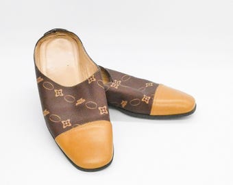 May include: A pair of brown slip-on mules featuring a repeating tan floral and oval pattern across the upper fabric. The shoes have a rounded tan leather toe cap, a tan leather interior lining, and a low-profile black sole.