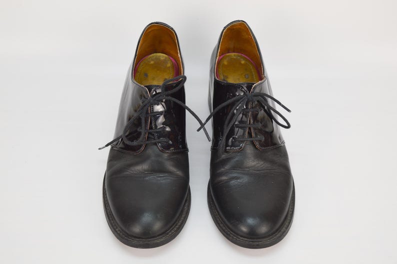 Puede incluir: Un par de zapatos negros de cuero con cordones y puntera de charol. Los zapatos tienen un estilo oxford cl&aacute;sico.