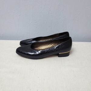 May include: A pair of black low-heeled pumps featuring a textured, reptilian-inspired embossed pattern on the exterior. The shoes have a rounded toe, a gold-toned metallic accent at the base of the heel, and a metallic gold-colored interior lining.