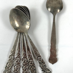 May include: A set of antique silver spoons. Six spoons are stacked, revealing intricate floral patterns on the handles. One spoon is separate, with a simpler design. The spoons have a vintage, tarnished look, suggesting age and use.