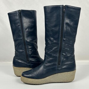 May include: A pair of navy blue tall boots with side zippers and beige wedge soles. The boots have a smooth leather-like texture and a vintage style. The zippers run along the side of each boot.