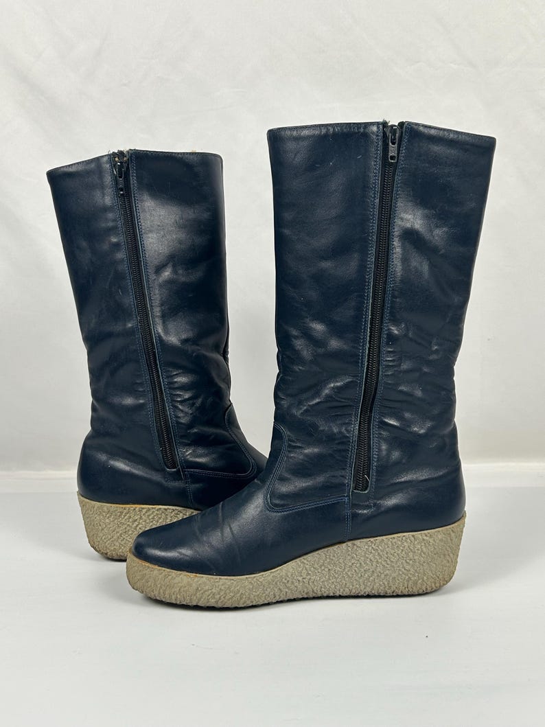 May include: A pair of navy blue tall boots with side zippers and beige wedge soles. The boots have a smooth leather-like texture and a vintage style. The zippers run along the side of each boot.