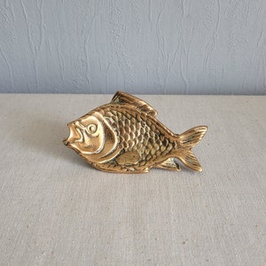 Puede incluir: Un plato de metal dorado en forma de pez. El pez tiene un diseño detallado con escamas y una boca ligeramente abierta.